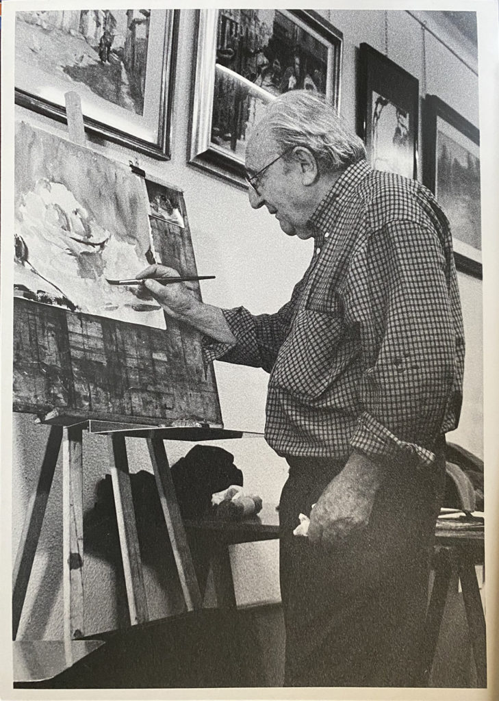 Luis Borruel painting a watercolour at his easel, framed works hanging behind him
