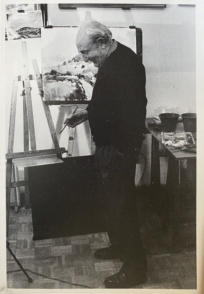 Luis Borruel at the easel in his studio, brush in hand, water pots and palette on the table beside him