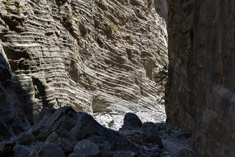 Sunlight on the layered limestone wall of the Samaria gorge, dark boulders and a dry white riverbed below