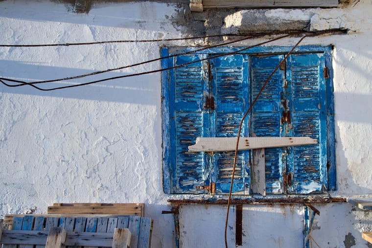 A blue louvred shutter flaking against a whitewashed wall, rusted wires strung across it