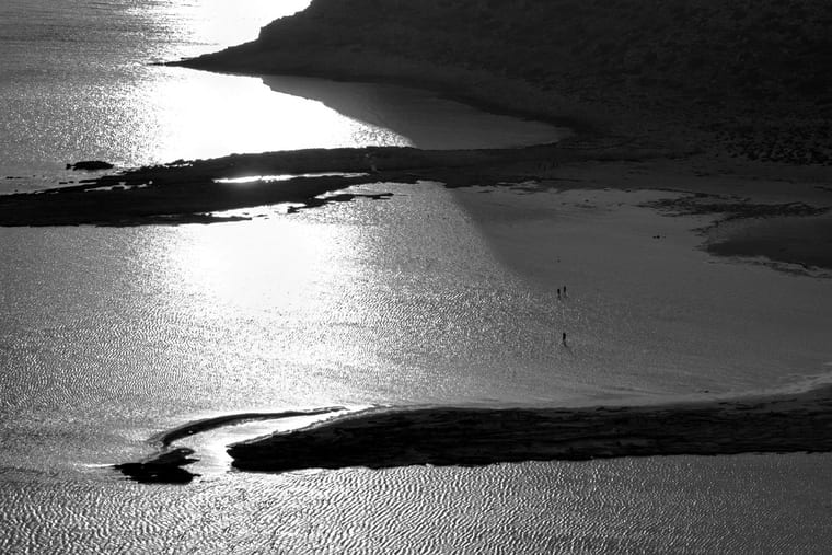 Sunlight glittering across the Balos lagoon seen from above, two walkers tiny on the sandbar