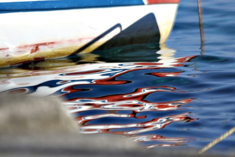 The hull of a moored boat and its red, white and blue reflection breaking up in the ripples