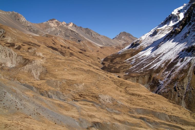 A dry ochre valley climbing between scree slopes to a snow-streaked ridge, under a cloudless deep blue sky