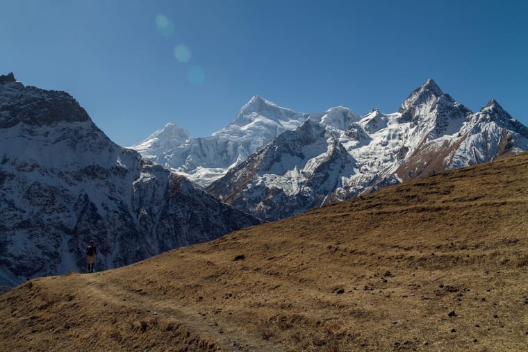 A lone walker on a path across a tawny hillside, dwarfed by the wall of snow peaks behind